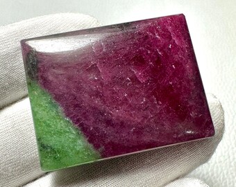 May include: A polished, rectangular gemstone with a vibrant color palette. The stone features a deep magenta hue dominating the surface, with a diagonal section of bright green. The gemstone is held by a gloved hand.