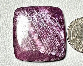 May include: A square-cut, polished gemstone with a deep purple hue and a shimmering, textured surface. The stone's facets catch the light, creating a play of color. A US quarter coin is next to the gemstone for size comparison.