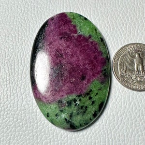 May include: Oval-shaped gemstone with a polished surface. The stone features a vibrant mix of colors, including deep red, green, and black. A US quarter coin is next to the gemstone for size comparison. The gemstone is likely a ruby in zoisite.