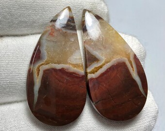 May include: Two teardrop-shaped cabochons of red, brown, and white banded agate. The stones are polished and have a smooth, glossy finish.
