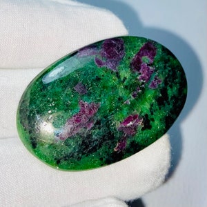 Può includere: Pietra preziosa di forma ovale, con una superficie lucida che mostra una sorprendente combinazione di verde e rosso intenso. Il motivo della pietra è maculato, con gruppi irregolari di rosso incastonati in una base verde. La gemma è presentata su uno sfondo bianco.