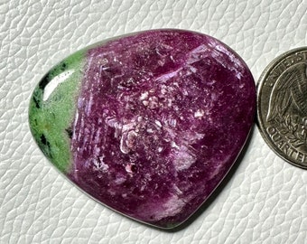 May include: A polished ruby zoisite gemstone with a heart-like shape. The stone features a deep magenta hue with green and black accents. A US quarter coin is next to the gemstone for size comparison.