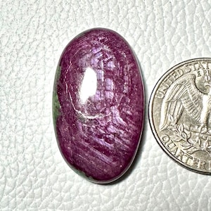 Puede incluir: Gema ovalada de color burdeos intenso, que exhibe una textura fibrosa y brillante. Una moneda de cuarto de dólar de plata se coloca junto a la gema para comparar el tamaño. La gema tiene una superficie pulida y un sutil borde verde.