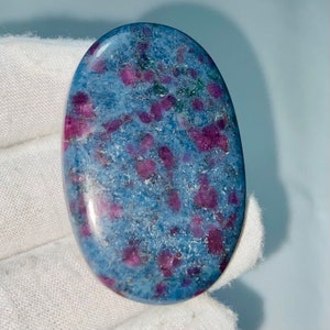 May include: Oval-shaped gemstone with a light blue base speckled with vibrant, deep red and green inclusions. The polished stone has a smooth, glossy surface, showcasing the natural patterns and colors of the mineral.