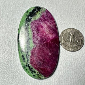 Può includere: Una gemma ovale con un vivace mix di colori. La pietra presenta una sorprendente combinazione di magenta intenso, verde e nero. Una moneta è posta accanto alla gemma per il confronto delle dimensioni.