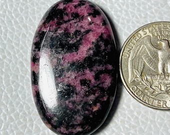 Puede incluir: Una piedra preciosa ovalada con superficie pulida. La piedra presenta una mezcla de colores negro y rosa intenso. Una moneda se coloca junto a la piedra preciosa para comparar el tamaño.