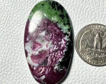 May include: Oval-shaped ruby zoisite gemstone with a quarter coin for size comparison. The stone features a mix of colors, including deep red, green, white, and black. The polished surface reflects light, highlighting the natural patterns and textures.