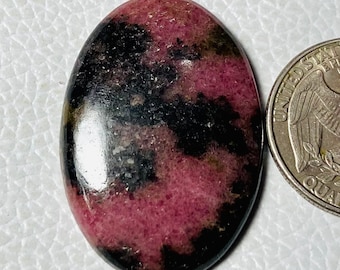 May include: Oval-shaped gemstone with a mix of pink and black colors. The polished stone has a smooth surface and is next to a quarter coin for size comparison. The gemstone's pattern is a mix of dark and light shades.