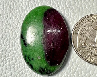 May include: An oval-shaped polished zoisite gemstone featuring a vibrant green base with a large, deep purple-red inclusion and small black speckles, positioned next to a United States quarter coin for scale to show its relative size.