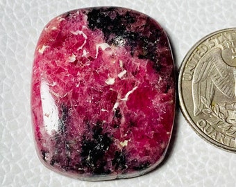 Puede incluir: Una gema pulida, rectangular, con un tono rosa vibrante, intercalada con inclusiones negras y blancas. La gema se muestra junto a una moneda para comparar el tamaño. La piedra tiene una superficie lisa y brillante.