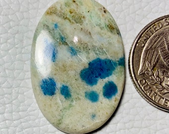 May include: Oval-shaped gemstone with a polished surface. The stone features a creamy white base color with blue and green inclusions. A US quarter coin is next to the stone for size comparison. The gemstone is likely for jewelry making.