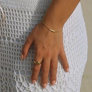 May include: A gold-colored bracelet with a small, elongated charm and a matching ring with a seashell design. The jewelry is worn on a hand, with a white crocheted garment in the background.