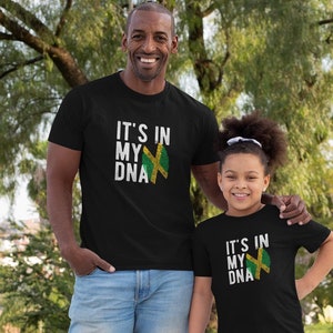May include: A father and child wearing matching black t-shirts with the text "It's in my DNA" and a Jamaican flag graphic. The shirts are a black color with white text and a green, yellow, and black flag graphic.