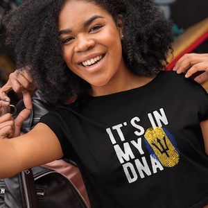 May include: Two people, one wearing a black crop top with the text "It's in my DNA" and a yellow, blue, and black flag design, and the other wearing a black leather jacket, are smiling and making a heart shape with their hands.