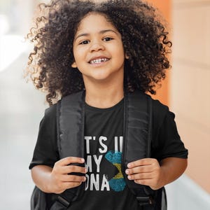 May include: A young person wearing a black t-shirt with a white graphic print of a fingerprint with the colors of the Bahamian flag. The text "It's in my DNA" is printed on the shirt. The person is smiling and wearing a black backpack.