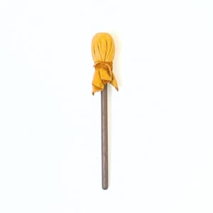Op de afbeelding: Een handgemaakte houten toverstaf met een gele stoffen kop. De toverstaf heeft een lange, slanke, bruine schacht en een afgeronde top gewikkeld in een geknoopte gele doek. De achtergrond is effen wit.