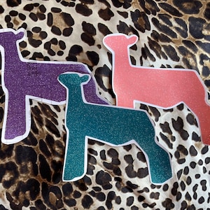 May include: Three glitter sheep silhouettes in purple, teal, and pink on a leopard print background.