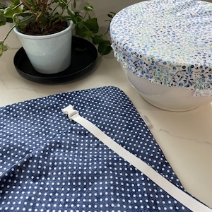 May include: Reusable bowl covers and a wrap. The bowl cover is white with a blue floral pattern and a ruffled edge. The wrap is navy blue with white polka dots and a white strap with a buckle.