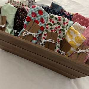 May include: A wooden tray filled with various fabric bowl covers. The covers feature colorful patterns including strawberries, cherries, and floral designs. Each cover is tied with twine and has a small tag that reads "Bowl Cover".