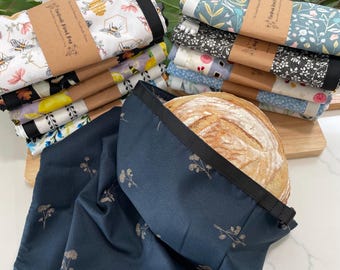 May include: A collection of fabric bread bags featuring various floral, bee, and botanical patterns, each secured with a brown paper band labeled Handmade Bread Bag. One dark blue bag with a floral print holds a round loaf of crusty sourdough bread.