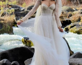 Puede incluir: Una novia con un vestido de novia de encaje blanco con un velo largo. El vestido tiene un corpiño ajustado y una falda fluida. La novia está de pie en un entorno natural con un río en el fondo.