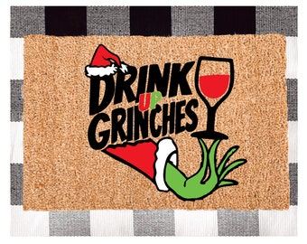 May include: A brown doormat with a black and white chequered border. The doormat features a green Grinch hand holding a glass of red wine. The text "Drink Up Grinches" is written in black letters.