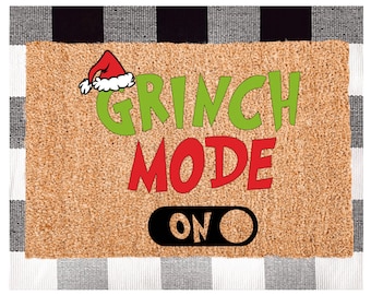 May include: A brown doormat with the words "Grinch Mode On" in red, green, and black lettering. A red Santa hat with white trim is above the word "Grinch".