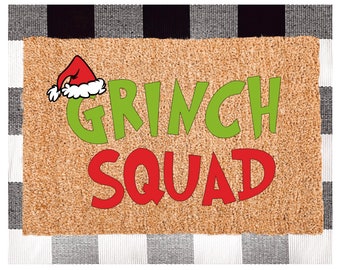 May include: A brown doormat with the words "Grinch Squad" in green and red letters. A red Santa hat with white trim is in the upper left corner.