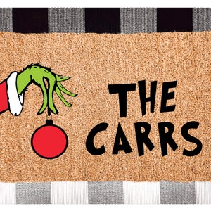 May include: A brown doormat with a green hand holding a red Christmas bauble. The text "THE CARRS" is printed in black on the doormat.