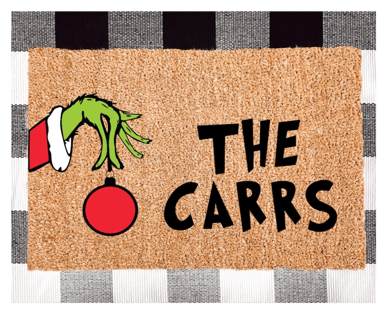 May include: A brown doormat with a green hand holding a red Christmas bauble. The text "THE CARRS" is printed in black on the doormat.