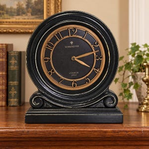May include: A round black table clock with a distressed finish, featuring a gold-toned face with black Arabic numerals. The center of the clock displays the text Kensington London 1892. It has two gold-colored hands and a sturdy rectangular base.
