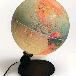 Op de afbeelding: Een vintage verlichte wereldbol met een zwarte voet en een netsnoer. De wereldbol toont een gedetailleerde kaart met oranje, groene en blauwe kleuren. De tekst "SCAN GLOBE DENMARK" is zichtbaar. De wereldbol is van binnenuit verlicht.
