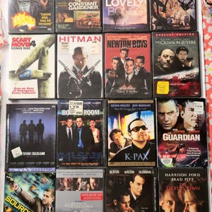Pode incluir: Uma coleção de DVDs com vários títulos de filmes, incluindo "Source Code", "Hitman" e "The Lovely Bones". As capas exibem atores e títulos de filmes, alguns DVDs têm extras. Os DVDs estão dispostos em uma superfície estampada.