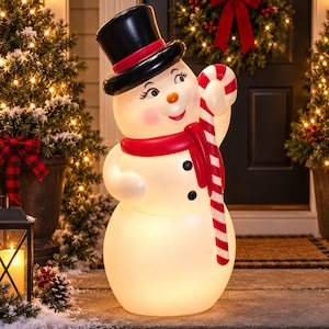 Pode incluir: Uma figura de boneco de neve branco com uma cartola preta com faixa vermelha, um cachecol vermelho e dois botões pretos no tronco. O boneco de neve tem um rosto sorridente com bochechas rosadas e segura uma grande bengala doce listrada.