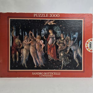 Op de afbeelding: Een puzzel van 2000 stukjes met Sandro Botticelli's kunstwerk "La Primavera". De afbeelding toont figuren in vloeiende gewaden tegen een donkere, beboste achtergrond. De puzzeldoos is rood met de tekst "PUZZLE 2000" en de naam van de kunstenaar.