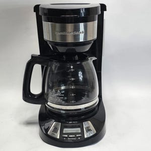 May include: A black Hamilton Beach coffee maker with a glass carafe. The carafe features white markings for 4, 6, 8, 10, and 12 cups. The base includes a digital display screen and several control buttons for programming brew options and settings.