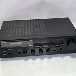 Könnte beinhalten: Schwarzer Yamaha Natural Sound Stereo-Receiver. Die Frontplatte verfügt über eine Digitalanzeige, mehrere Knöpfe und Tasten für verschiedene Funktionen. Der Receiver hat einen großen Lautstärkeregler auf der rechten Seite.