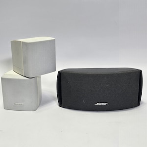 Puede incluir: Tres altavoces: dos altavoces cúbicos blancos y un altavoz negro curvado. Los altavoces blancos están apilados, con una parte frontal de malla. El altavoz negro tiene la marca "BOSE" en blanco en la parte delantera.
