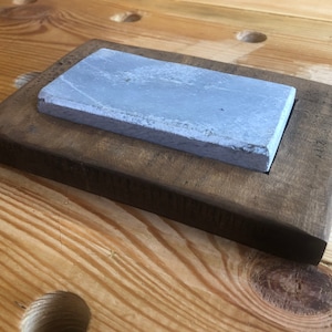 May include: A grey rectangular sharpening stone sits in a wooden frame. The frame is dark brown and has a slightly rounded edge.