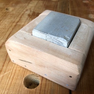 Pu&ograve; includere: Una pietra per affilare grigia che riposa in un supporto di legno. Il supporto &egrave; in legno chiaro e ha una forma rettangolare.