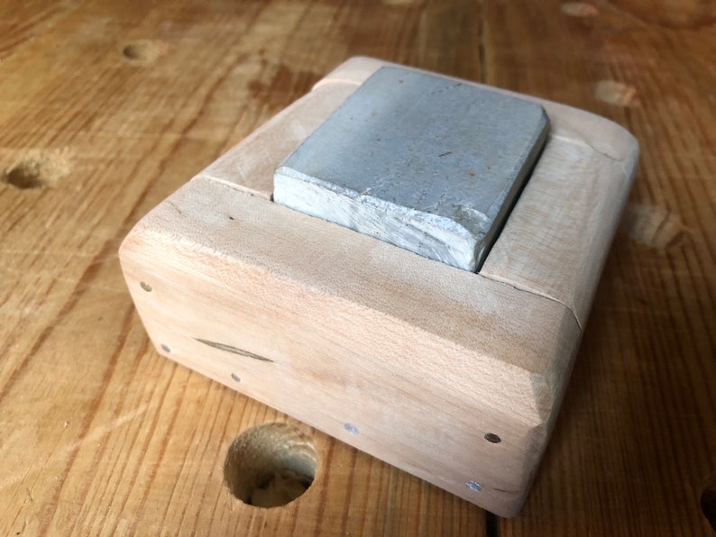 Pu&ograve; includere: Una pietra per affilare grigia che riposa in un supporto di legno. Il supporto &egrave; in legno chiaro e ha una forma rettangolare.