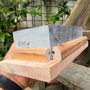 May include: A rectangular gray stone block with the text 1715 gr written on its side, secured within a light brown wooden frame. The frame consists of a flat base and two parallel wooden rails that hold the stone in place, showing wood grain.