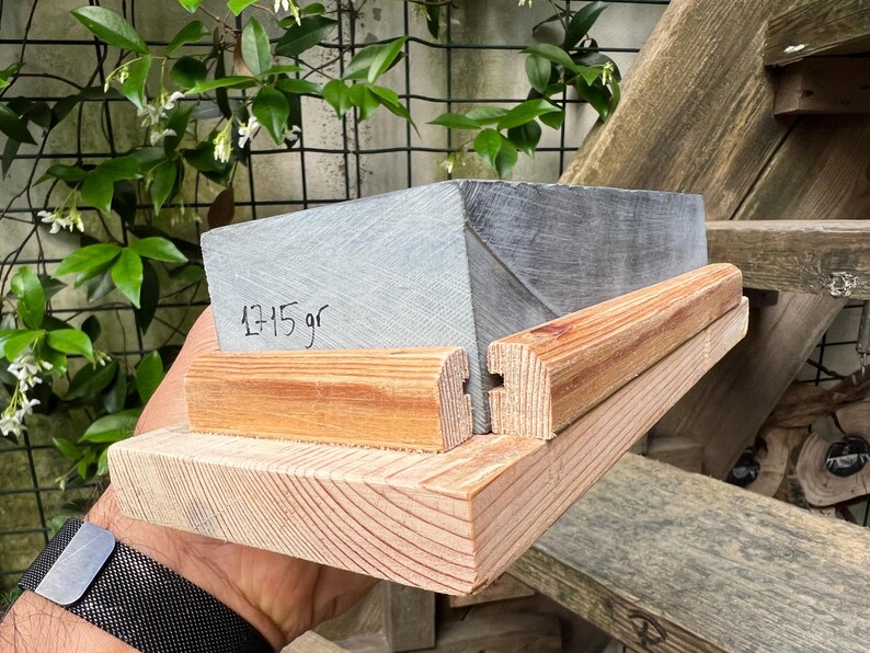 May include: A rectangular gray stone block with the text 1715 gr written on its side, secured within a light brown wooden frame. The frame consists of a flat base and two parallel wooden rails that hold the stone in place, showing wood grain.