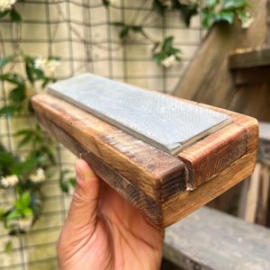 Può includere: Un ceppo per affilare coltelli in legno con una pietra rettangolare grigia. Il ceppo è realizzato in legno marrone chiaro e scuro. La pietra è inserita nel ceppo di legno. L'articolo è tenuto in una mano.