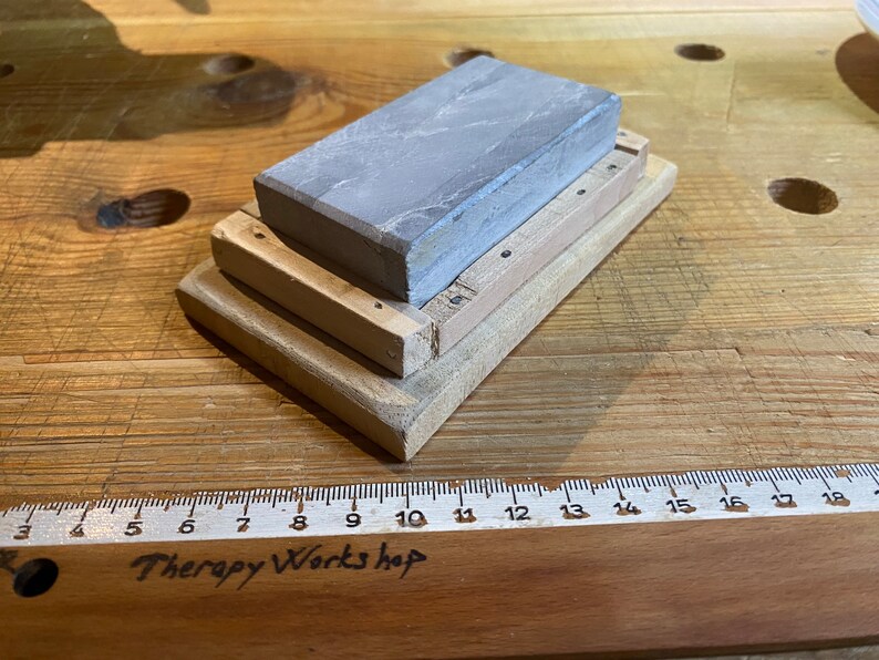 Peut inclure: Une pierre &agrave; aiguiser grise avec une base en bois. La base est compos&eacute;e de trois couches de bois, chacune avec un motif de grain diff&eacute;rent. La pierre mesure environ 15 cm de long et 5 cm de large.