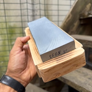 Puede incluir: Una piedra de afilar rectangular de color gris colocada en una base de madera clara. El lateral de la piedra presenta el texto escrito a mano 1665 gr, que indica su peso en gramos. La piedra tiene una superficie lisa y plana con textura.