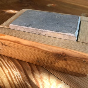 Puede incluir: Una peque&ntilde;a caja de madera con una piedra de pizarra gris en su interior. La caja est&aacute; hecha de madera de corte &aacute;spero y tiene un acabado natural.