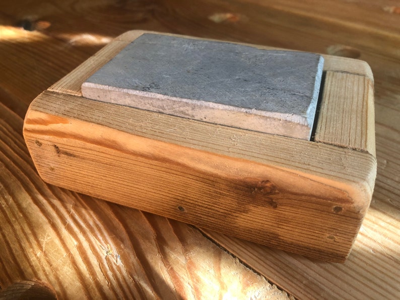 Puede incluir: Una peque&ntilde;a caja de madera con una piedra de pizarra gris en su interior. La caja est&aacute; hecha de madera de corte &aacute;spero y tiene un acabado natural.