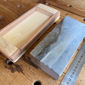 Puede incluir: Una caja de madera con un interior de madera clara y un exterior de madera oscura. La caja est&aacute; vac&iacute;a. Junto a la caja hay una piedra de afilar rectangular gris. Una regla de metal es parcialmente visible en el fondo.