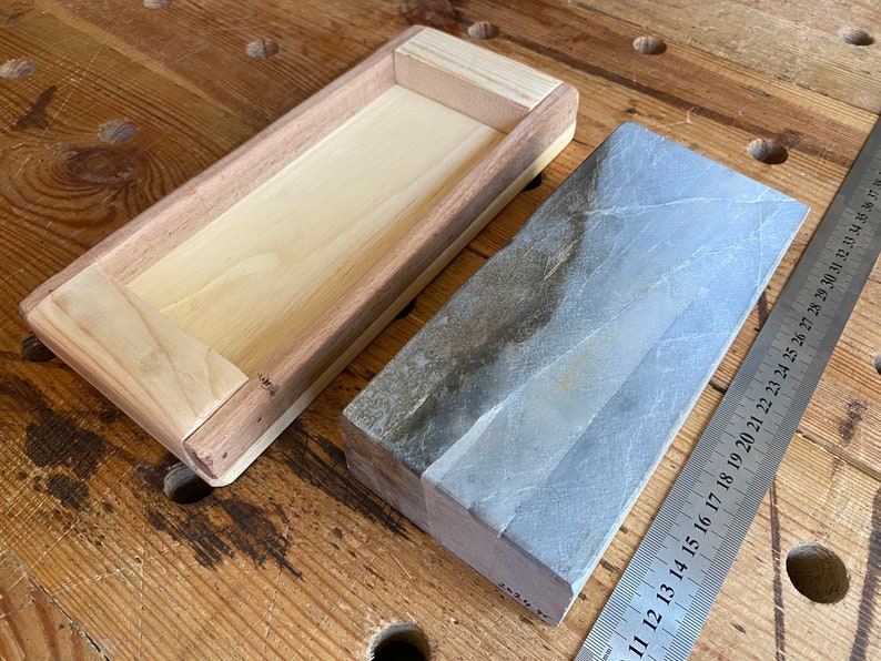 Puede incluir: Una caja de madera con un interior de madera clara y un exterior de madera oscura. La caja est&aacute; vac&iacute;a. Junto a la caja hay una piedra de afilar rectangular gris. Una regla de metal es parcialmente visible en el fondo.